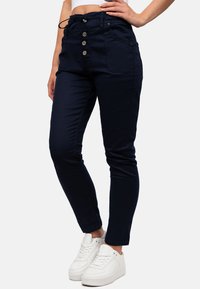 Pantalon bleu marine avec taille à cordon, comportant trois boutons à l'avant, des poches latérales et une coupe fuselée. Porté avec des baskets blanches.