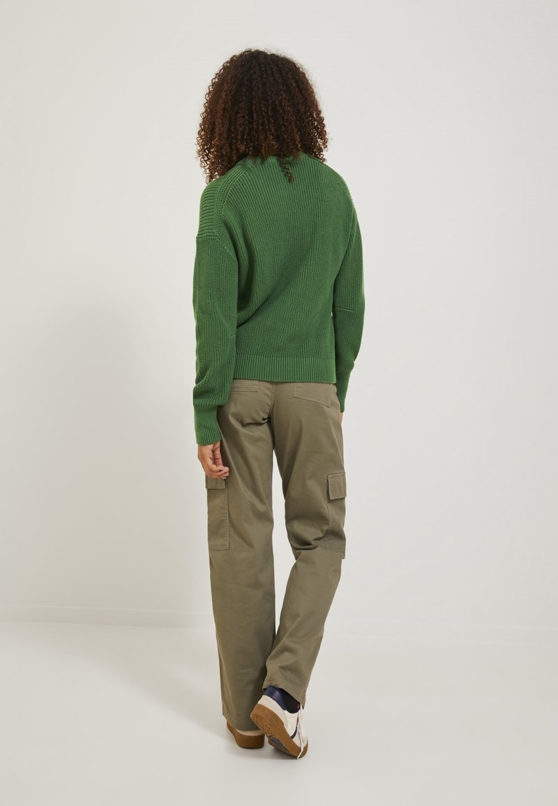 Personne aux cheveux bouclés portant un pull en maille vert, un pantalon cargo kaki et des baskets blanches, marchant de dos sur un fond uni.