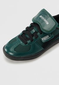 Zapatilla deportiva de color verde oscuro con parte superior de cuero sintético liso, cordones negros y una correa de velcro con el texto "Slytherin". Suela de goma negra.