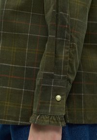 Gros plan d'une manche de chemise en velours côtelé à carreaux verts avec un poignet boutonné or sur un jean bleu en denim.