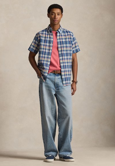 Άνδρας που στέκεται, φοράει μπλε καρό πουκάμισο με κοντά μανίκια πάνω από κόκκινη μπλούζα, ανοιχτό μπλε τζιν με φαρδιά μπατζάκια, ναυτικό μπλε αθλητικά παπούτσια, καφέ ζώνη και ρολόι.