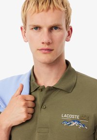 Giovane con i capelli biondi corti che indossa una polo Lacoste verde oliva con ricamo di montagne, tenendo un capo di abbigliamento azzurro chiaro sulla spalla.