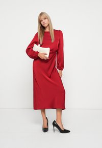 Robe midi en satin rouge avec de longues manches bouffantes, fente sur le côté, accompagnée de talons hauts noirs et d'une pochette blanche. Texture lisse sur l'ensemble.