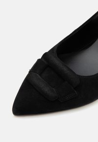 Chaussure noire en daim à bout pointu avec une grande boucle assortie en daim sur le dessus, présentant une doublure intérieure lisse.