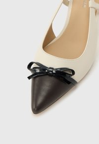 Γυναικεία δερμάτινη μπαλαρίνα διχρωμία σε κρεμ και μαύρο με μαύρο γυαλιστερό φιόγκο στο επάνω μέρος και λουράκι που πιασμένο πίσω από το τακούνι.