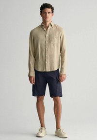Beige Leinenhemd, zugeknöpft, mit einer Brusttasche; navy Cargo-Shorts mit Seitentaschen; cremefarbene Sneakers aus strukturiertem Material, stehen vor einem neutralen Hintergrund.