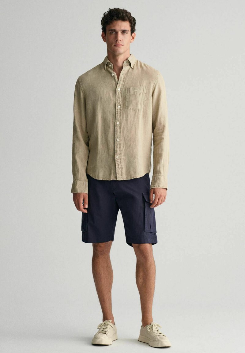 Beige Leinenhemd, zugeknöpft, mit einer Brusttasche; navy Cargo-Shorts mit Seitentaschen; cremefarbene Sneakers aus strukturiertem Material, stehen vor einem neutralen Hintergrund.