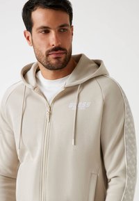 Beige zip-up hoodie met trekkoorden, voorzien van een wit logo op de borst en patroonaccenten langs de mouwen. Zacht, gestructureerd materiaal.