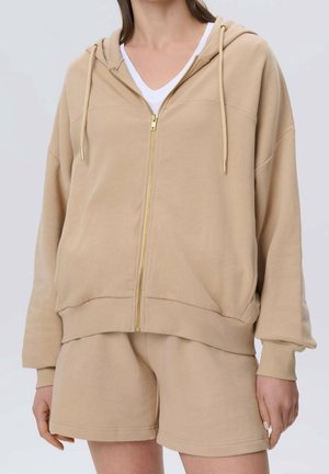 Personne portant un sweat à capuche beige zippé avec cordons et un short beige assorti, avec un t-shirt blanc visible en dessous.