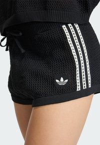 Čierne pletené šortky s bielym logom Adidas trefoil a tromi bielymi pruhmi na bokoch s malými čiernymi trefoilmi, nosené na nahých nohách.