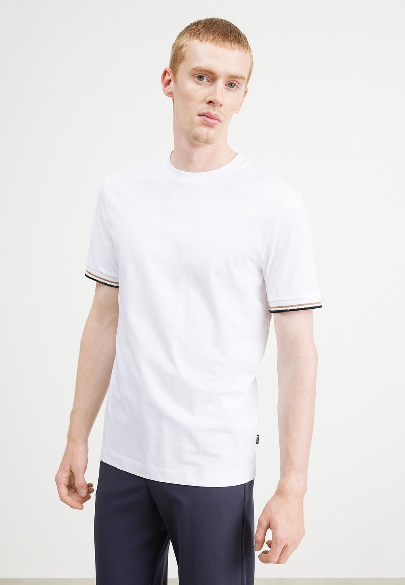 Weißes Baumwoll-T-Shirt mit kurzen Ärmeln, das braune und schwarze Streifenakzente, einen runden Ausschnitt und ein Etikett an der linken Seitennaht aufweist.