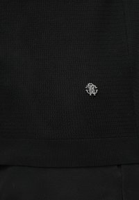 Tissu tricoté noir texturé avec un petit emblème logo métallique argenté près du bord central inférieur.