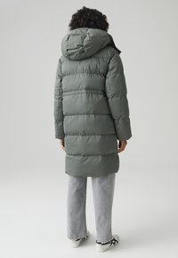 Lunga giacca puffa in verde tenue con cappuccio, con sezioni trapuntate e texture liscia, abbinata a pantaloni a gamba larga grigio chiaro e sneakers.