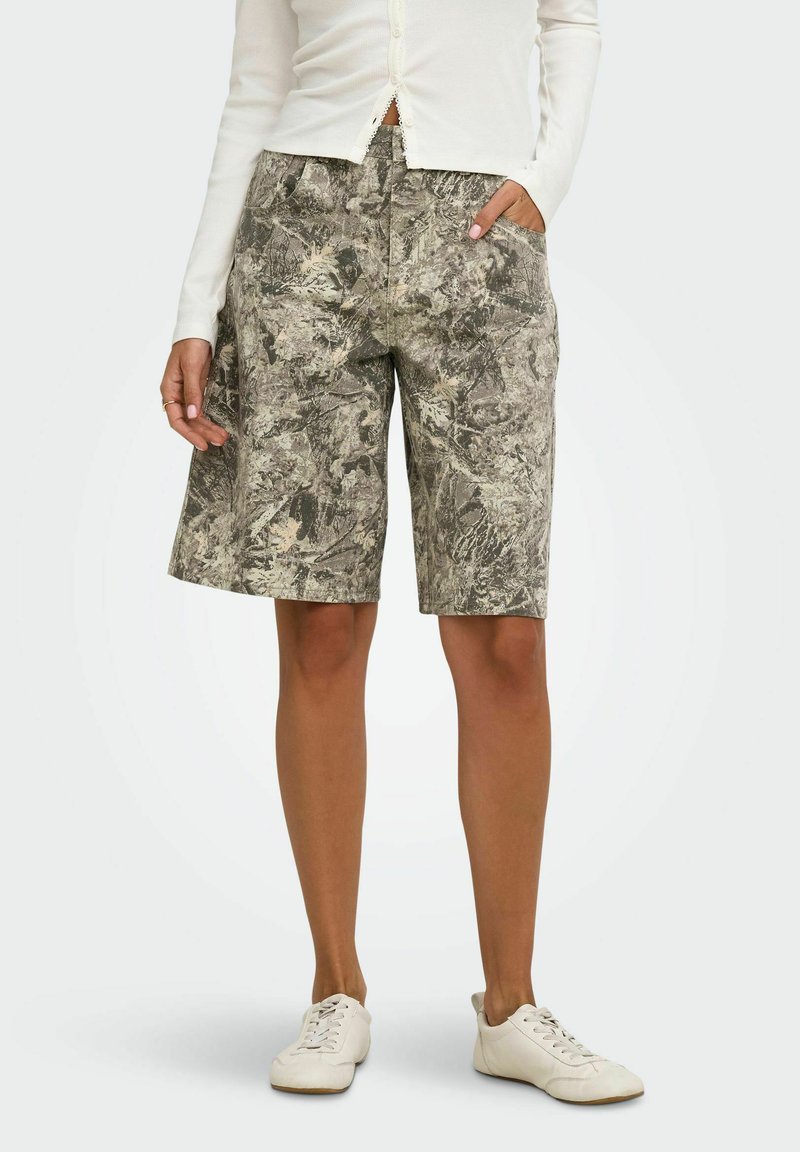 Femme portant un short gris à motifs longueur genou, un haut blanc à manches longues boutonné et des baskets blanches, debout avec une main dans la poche.