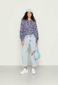 Mulher vestindo uma blusa floral, jeans claros rasgados, tênis brancos, chapéu de balde branco, segurando uma bolsa azul clara contra um fundo simples.