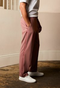 Rosa skræddersyede bukser i et glat stof med sidelommer, parret med hvide sneakers, set fra siden mod en neutral baggrund.