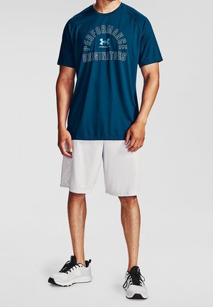 Homme portant une chemise bleue à manches courtes avec le texte "Performance Originators", un short blanc et des chaussures de sport blanches avec des lacets noirs.