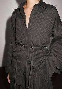A close-up of a person wearing a black, pinstriped shirt with a collar, featuring a tied waist and long sleeves, against a neutral background.