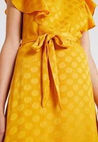 Gul klänning med en strukturerad tyg, med ett prickigt mönster och ett knytbart midjebälte. Volangärmar ger detaljer till designen.