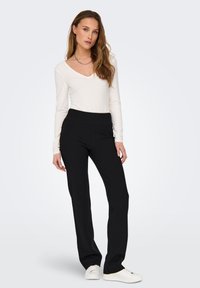 Sorte bukser med bred linning, slim-fit design og glat tekstur, parret med en hvid langærmet top og hvide sneakers.