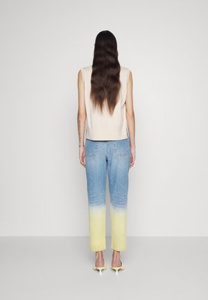 Vrouw met lang donker haar, zij draagt een mouwloze beige top, jeans met een blauw-naar-geel ombre-effect en gele sandalen met hakken, staand met haar gezicht naar een effen witte muur toe.