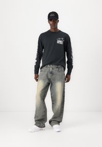 Grau langärmliges Hemd mit weißem Text und Grafiken, kombiniert mit ausgewaschenen, weit geschnittenen Jeans und weißen Turnschuhen. Schwarze Mütze mit Text.