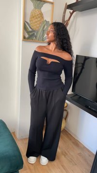 Top nero off-the-shoulder con un taglio, abbinato a pantaloni neri larghi. Modella che indossa sneakers bianche con platform, in piedi all'interno vicino a una TV.