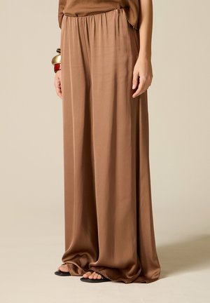 Losse bruine broek met elastische taille, gedragen door een persoon die staat, gecombineerd met zwarte sandalen met open teen en dikke armbanden aan één pols.