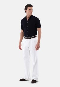 Polo shirt navy a maniche corte con bottoni, abbinato a pantaloni bianchi larghi e cintura marrone. Il modello è in piedi su uno sfondo neutro.