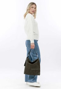 Dark green leather hobo bag with a slouchy design, zippered pocket, and a detachable strap. Model wears white sweater and light blue jeans.
