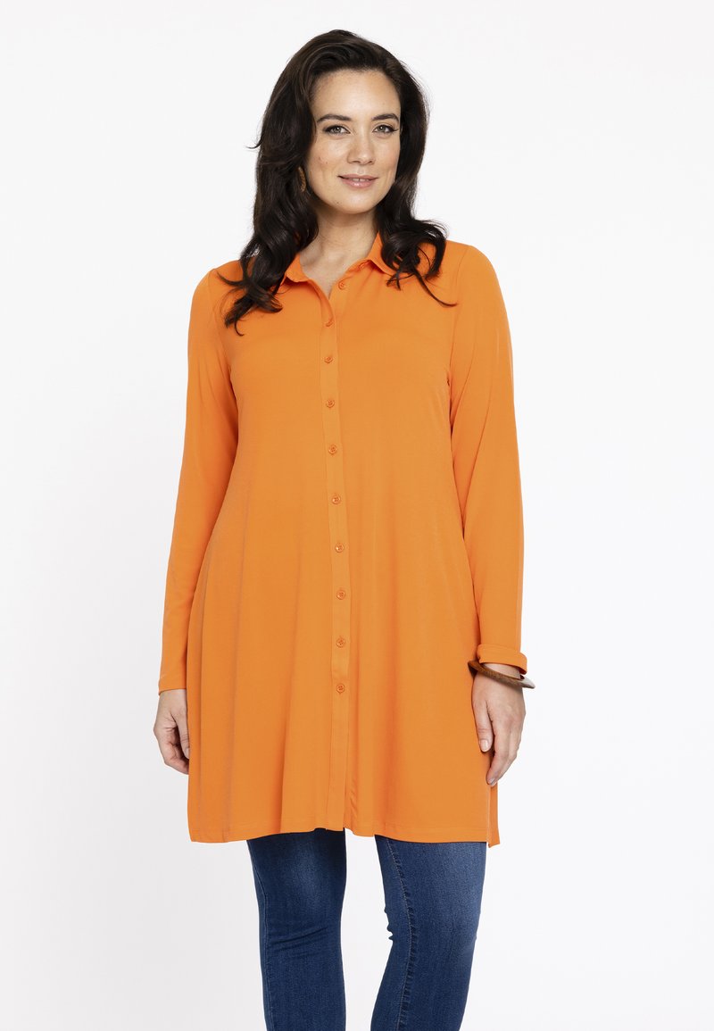 Orange langærmet skjorte med krave, lige snit og let buet nederkant, parret med blå skinny jeans.