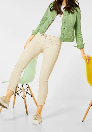 Jeans skinny beige con puños enrollados, chaqueta de mezclilla verde sobre una camiseta blanca, combinados con zapatillas ligeras, contra un fondo minimalista.