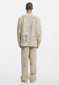 Set compus din jachetă și pantaloni bej, cu imprimeuri grafice albastre, având diverse modele inclusiv forme și simboluri; croială lejeră și material casual.