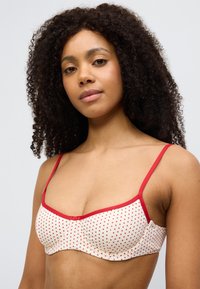 Σουτιέν με κρέμ βάση, κόκκινες πουά και αντίθετες κόκκινες τιράντες. Διαθέτει ελαφριά επένδυση και λεία υφή. Χωρίς επιπλ έον διακοσμητικά.