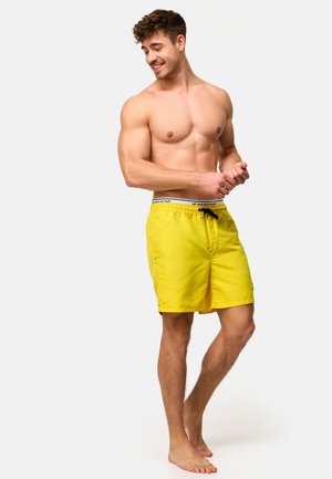 Muskeløs mand barfodet, smilende og kigger ned, iført lysegule badeshorts med sort snor på ensartet lys baggrund.