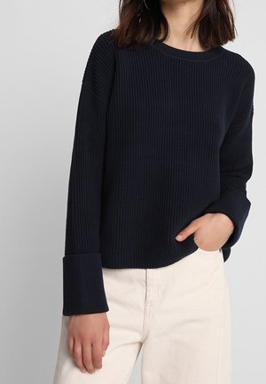Pull en maille côtelée bleu marine avec un col rond et des poignets larges. Le tissu semble texturé et épais, offrant une coupe décontractée.