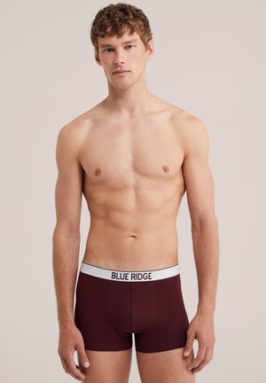 Giovane uomo con capelli ricci che indossa boxer rosso scuro con una cintura bianca con la scritta Blue Ridge, in piedi davanti a uno sfondo semplice.