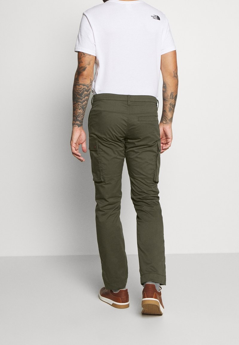 Homme aux bras tatoués portant une chemise blanche à manches courtes, un pantalon cargo vert olive et des baskets marron, vu de dos sur un fond uni.