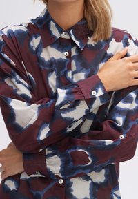 Chemise à manches longues dans un motif abstrait bordeaux, bleu et blanc. Présente un col classique et une fermeture boutonnière avec des boutons blancs.
