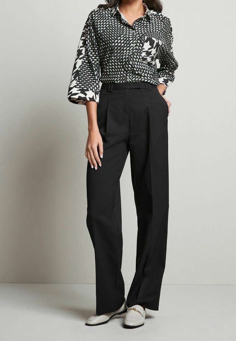 Black high-waisted trousers paired with a black and white patterned button-up shirt featuring various geometric designs and a chest pocket.