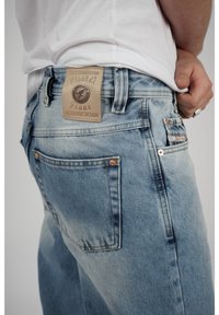Ljusblå denimjeans med en läderlogotyp på baksidan, kopparnitar och två bakfickor med subtila sömdetaljer.