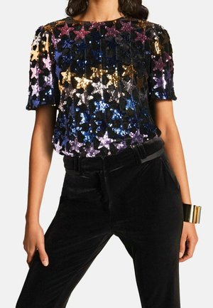 Femme portant un haut à sequins multicolore à motif d'étoiles, un pantalon en velours noir et un bracelet manchette doré à son poignet droit.