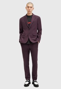 Un costume violet foncé composé d'un blazer ajusté et d'un pantalon slim, associé à un t-shirt graphique noir et des mocassins noirs avec des accents blancs.