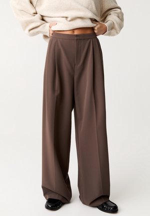 Person wearing wide-leg brown trousers, beige sweater lifted to show midriff, and black shoes on a plain light background.