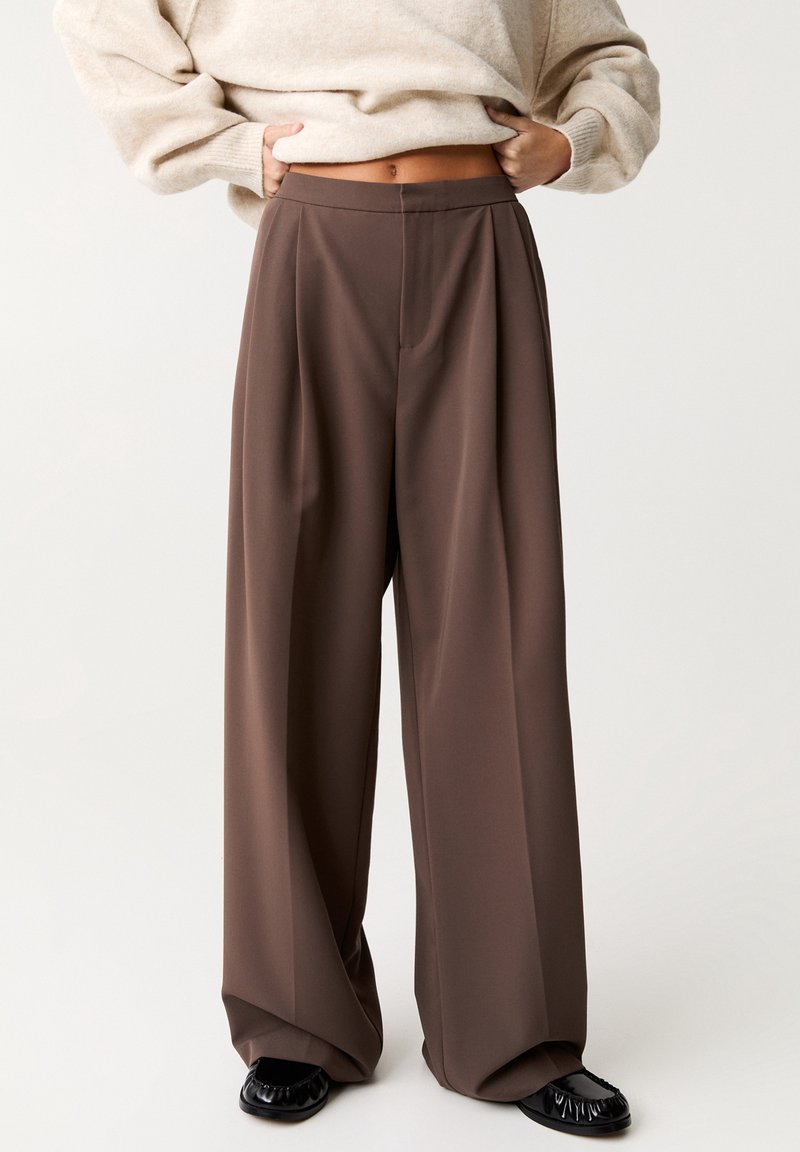 Personne portant un pantalon large marron, un pull beige relevé pour montrer le ventre, et des chaussures noires sur un fond clair uni.