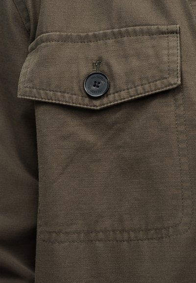 Τσέπη από ύφασμα σε ελαιώδες πράσινο με καπάκι ασφαλισμένο με μαύρο κουμπί που φέρει ανάγλυφη τη φράση "THE KOOPLES".