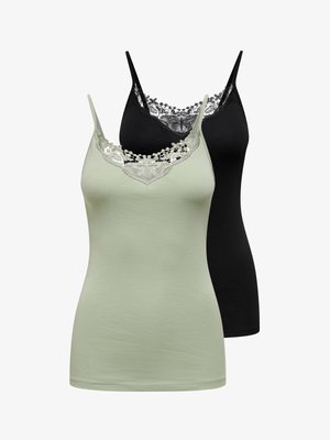 Lachende vrouw die een zwarte top met kanten rand draagt, een high-waisted versleten spijkershort en zwarte muiltjes met hak, staand tegen een witte achtergrond.
