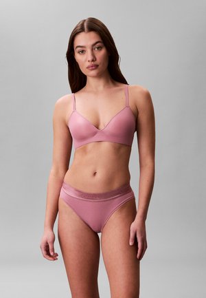 Femme portant un ensemble de sous-vêtements Calvin Klein assorti de couleur rose poussiéreux, debout sur un fond gris uni, faisant face à l'avant avec une expression neutre.