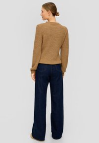 Brauner gerippter Strickpullover mit langen Ärmeln, kombiniert mit weit geschnittenen, dunkelblauen Jeans. Das Outfit zeichnet sich durch einen lässigen Schnitt und eine strukturierte Textur aus.