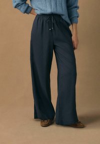 Pantaloni evazați bleumarin cu talie ajustabilă prin șnur și textură ușoară și fină, asortați cu un pulover albastru cu dungi și pantofi mocasini maro.